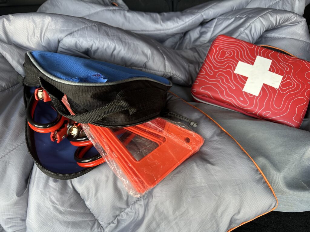 Image of jumper cables, bright orange hazard cones and a first aid kit in the back of a car with blankets.