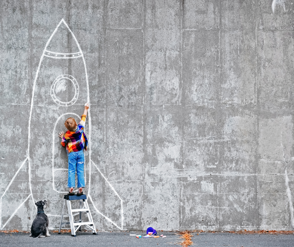 Photo of a child standing in front of a chalk drawn rocket ship. Dreaming can become reality and we should shoot for the stars.